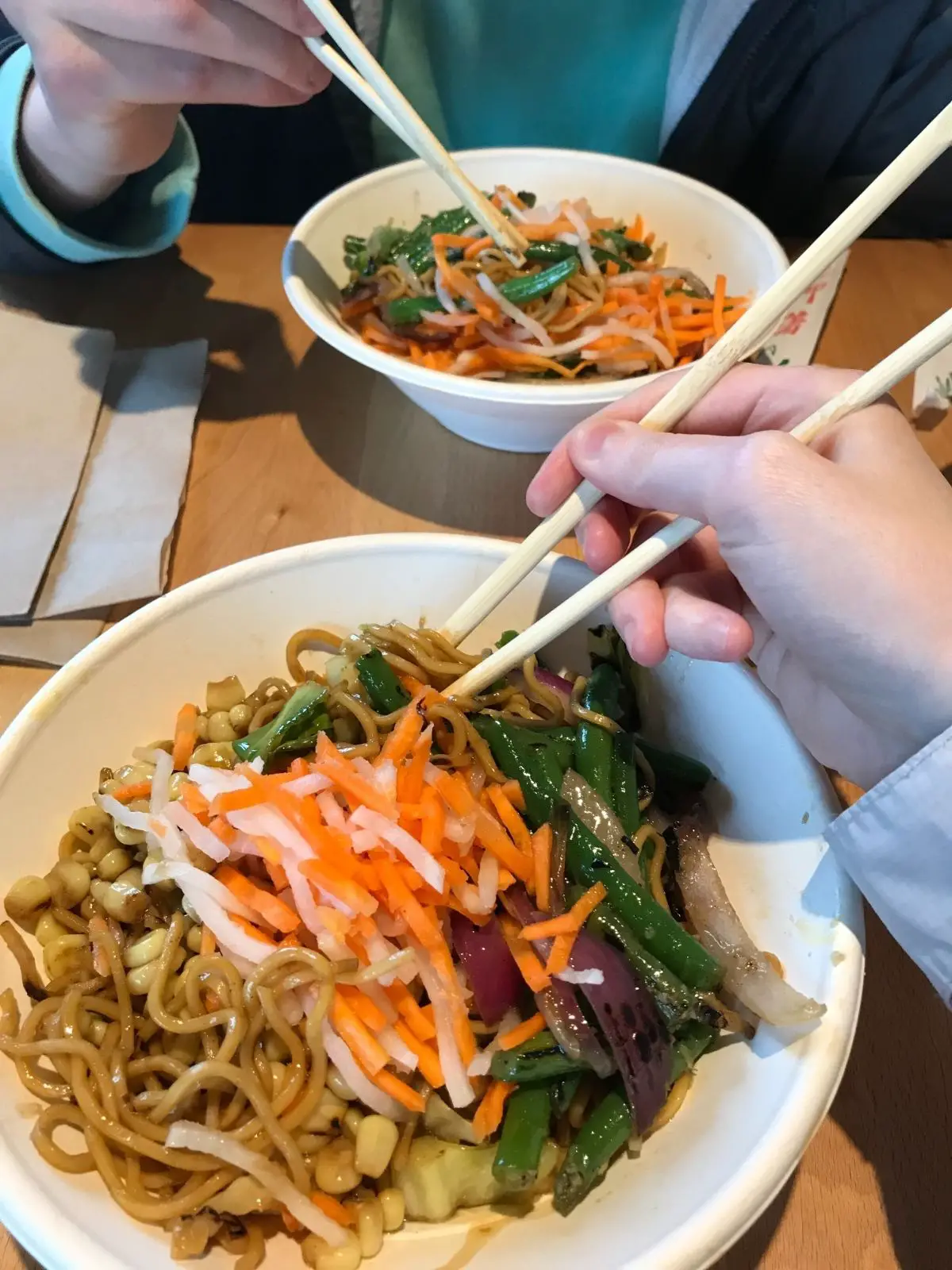 two bowls from zao that have yakisoba noodles, corn, green beans, carrots, and onions and are being eaten with chopsticks