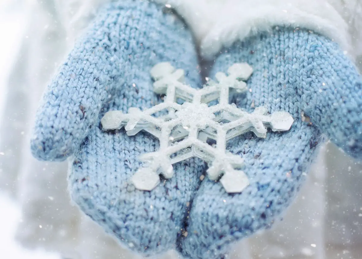 Someone wearing blue mittens is holding a huge snowflake while it snows.
