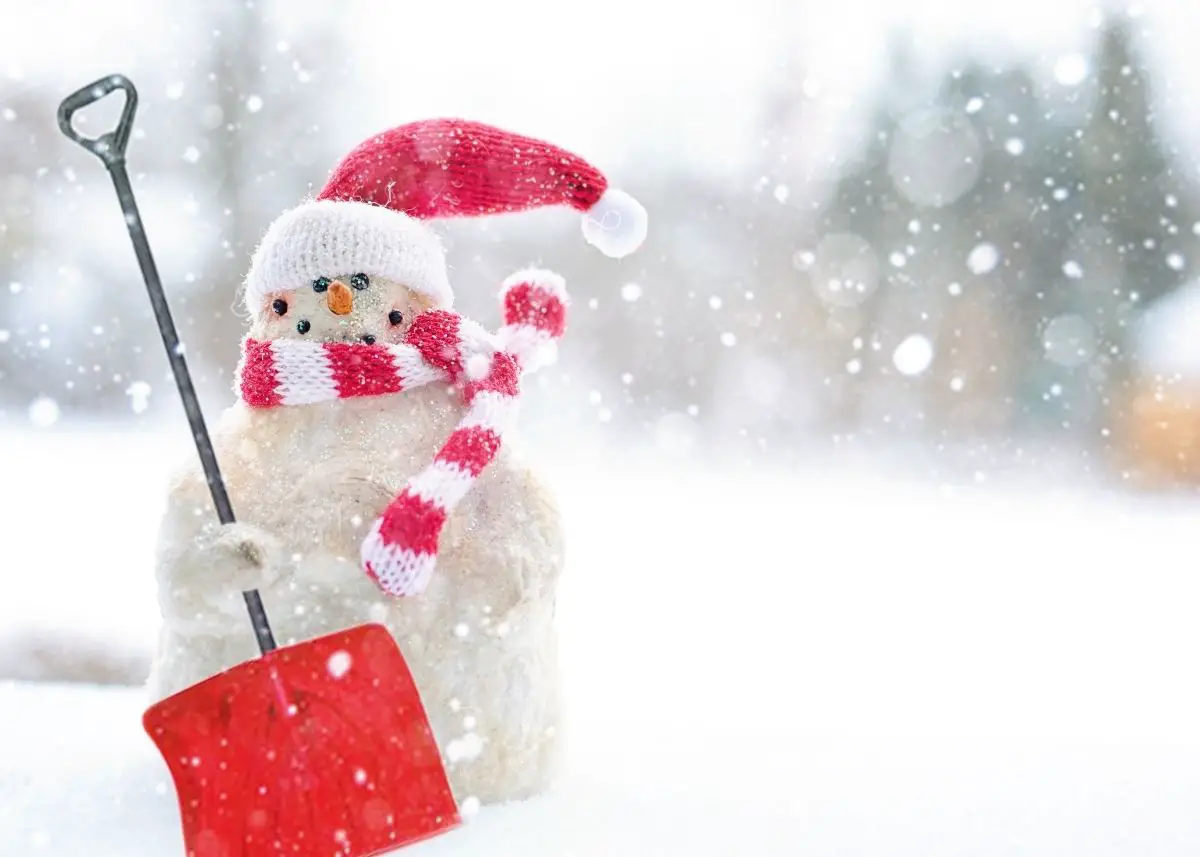 A happy snowman wearing a red and white hat and scarf is holding a red shovel.