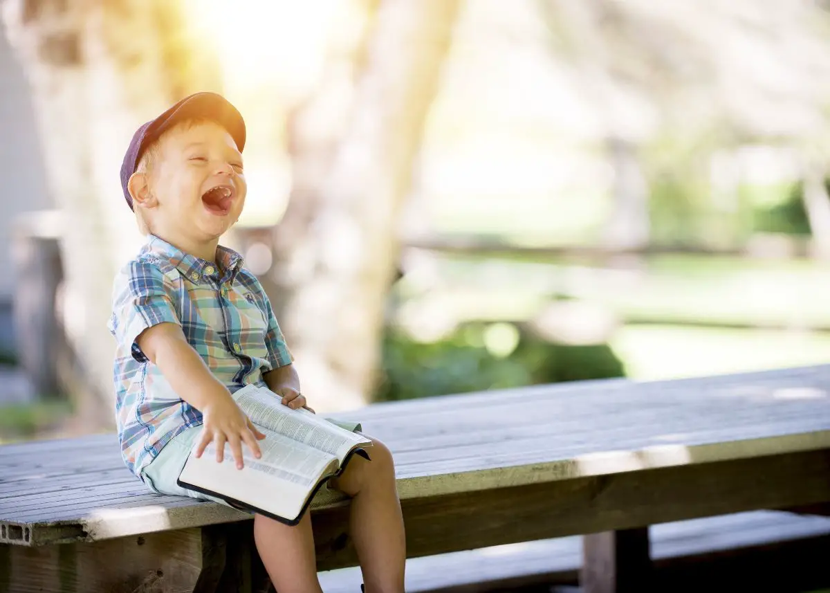 A little boy is sitting on a wood bench.  He's holding a book and laughing because he heard the best puns.