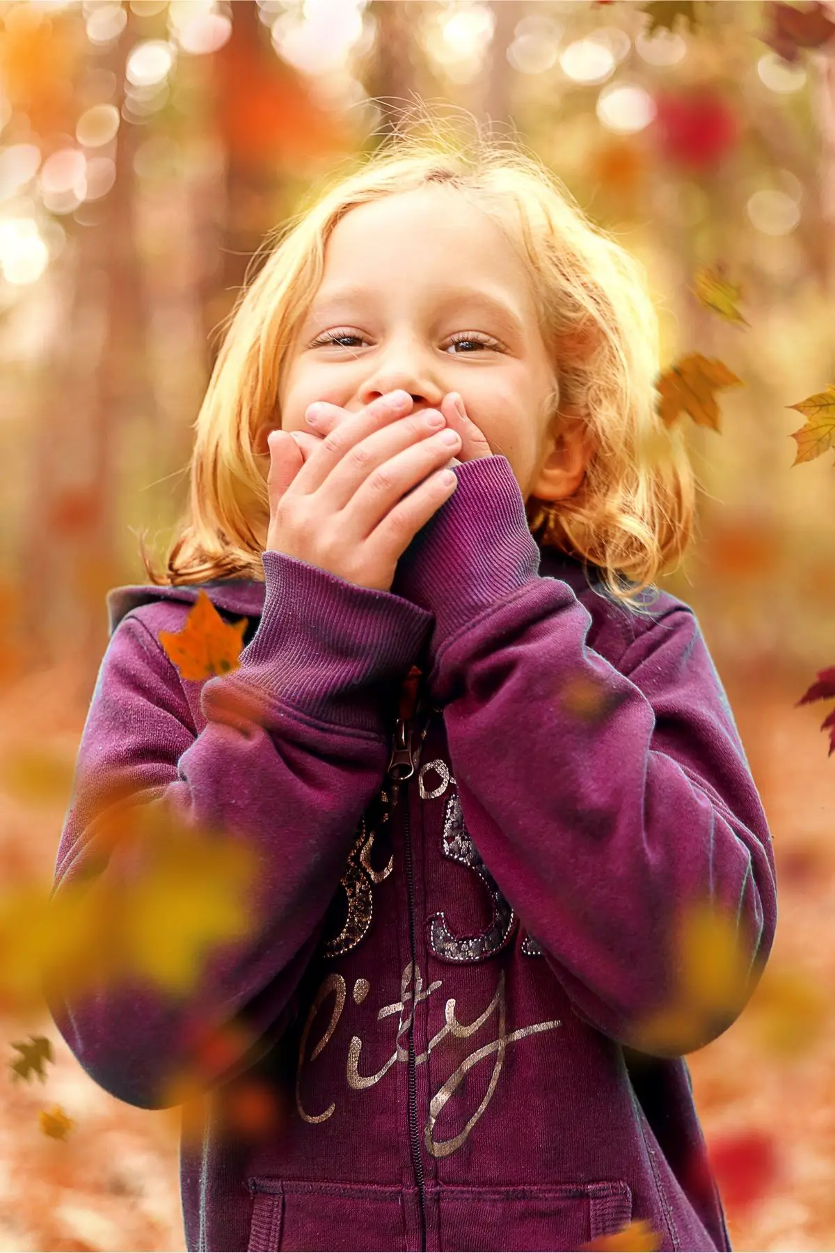 A blond haired girl wearing a purple sweatshirt is laughing.  She has her hands over her mouth and it's fall.
