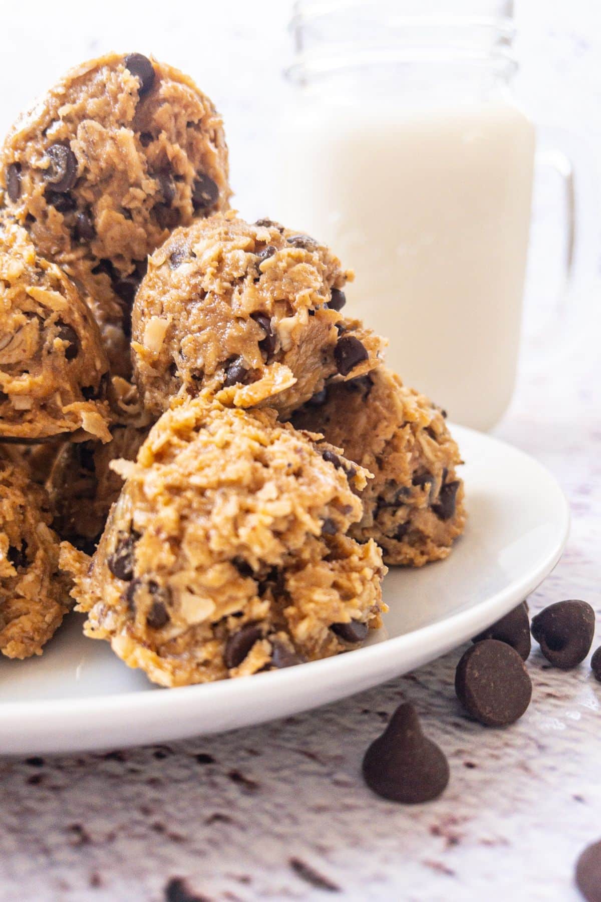 Protein balls stacked on a white plate with chocolate chips and a glass of milk.