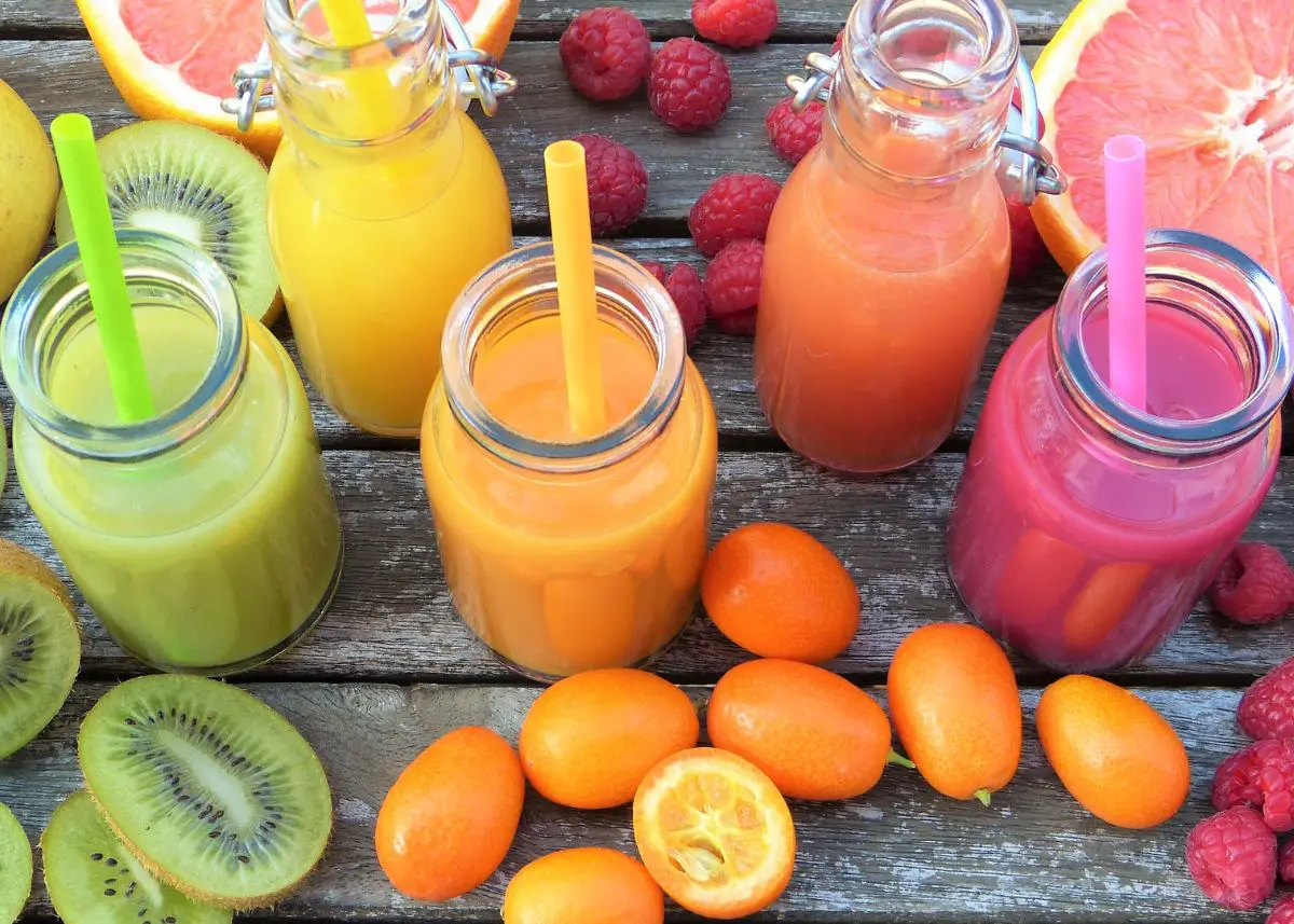 An overhead shot of five kinds of juice in different glass jars.  Four of the five have a straw.  They are surrounded by grapefruit, raspberries, oranges, and kiwis.