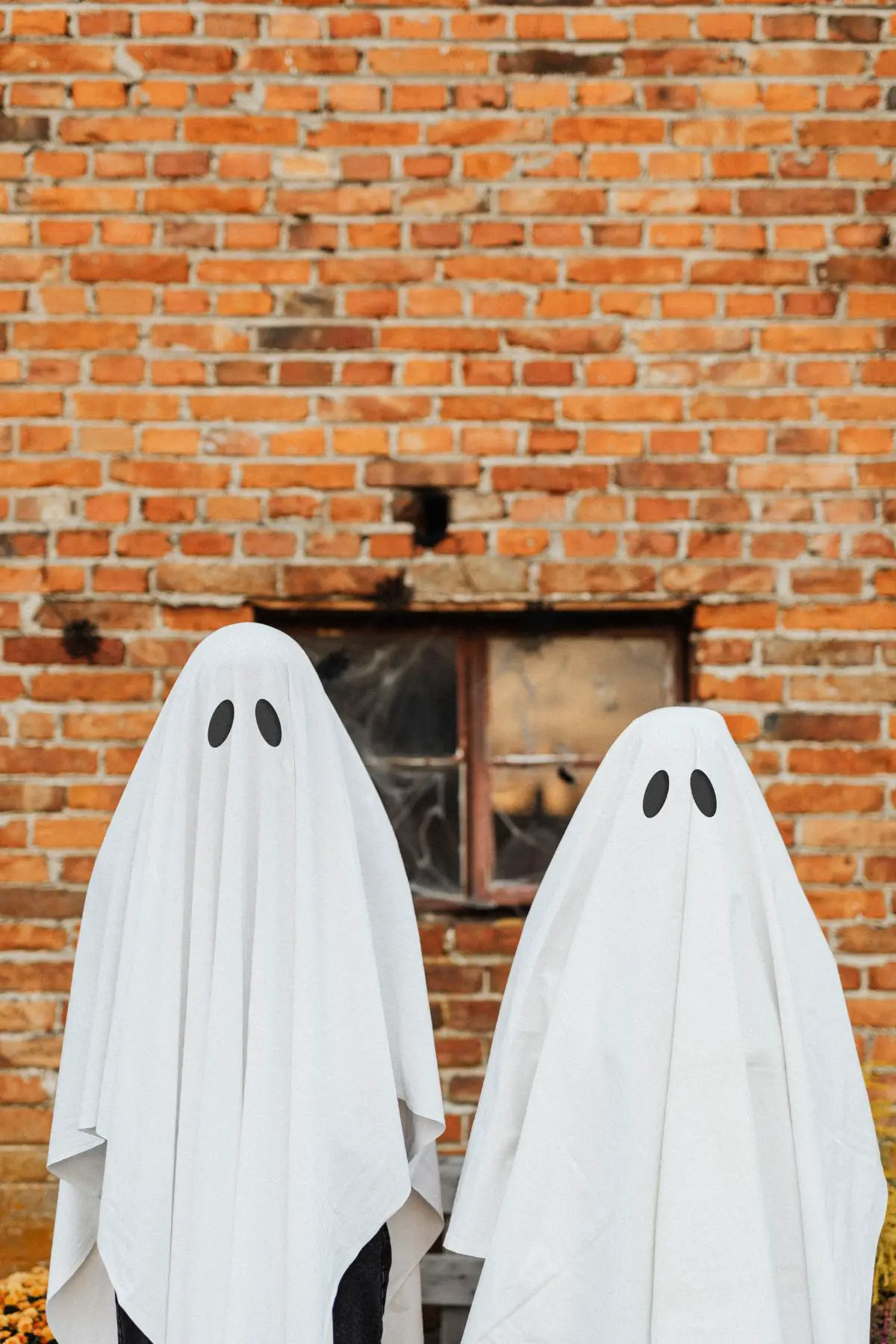 Two kids dressed as ghosts standing outside of a brick building.  