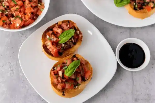Two slices of toasted french bread topped with bruschetta, balsamic glaze, and fresh basil leaves on a white plate with the bowl of bruschetta, bowl of balsamic glaze, and another plate of bruschetta bread in the background