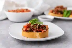 A slice of toasted french bread topped with bruschetta, balsamic glaze, and fresh basil leaves on a white plate with the bowl of bruschetta, bowl of balsamic glaze, and another plate of bruschetta bread in the background