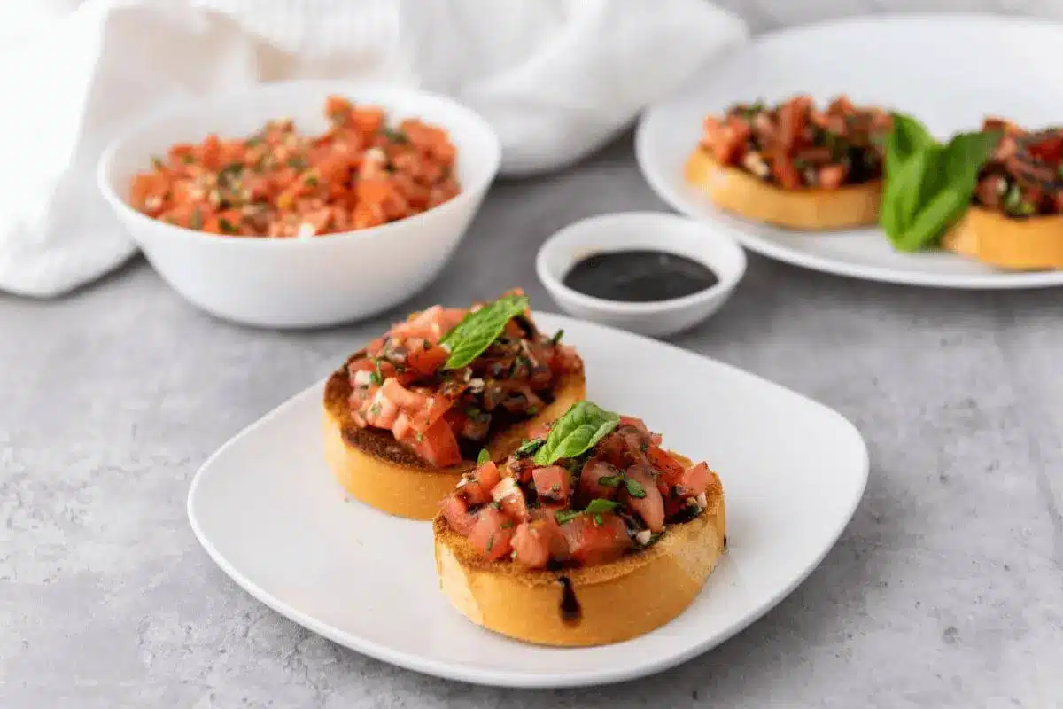 Two slices of toasted french bread topped with bruschetta, balsamic glaze, and fresh basil leaves on a white plate with the bowl of bruschetta, bowl of balsamic glaze, and another plate of bruschetta bread in the background