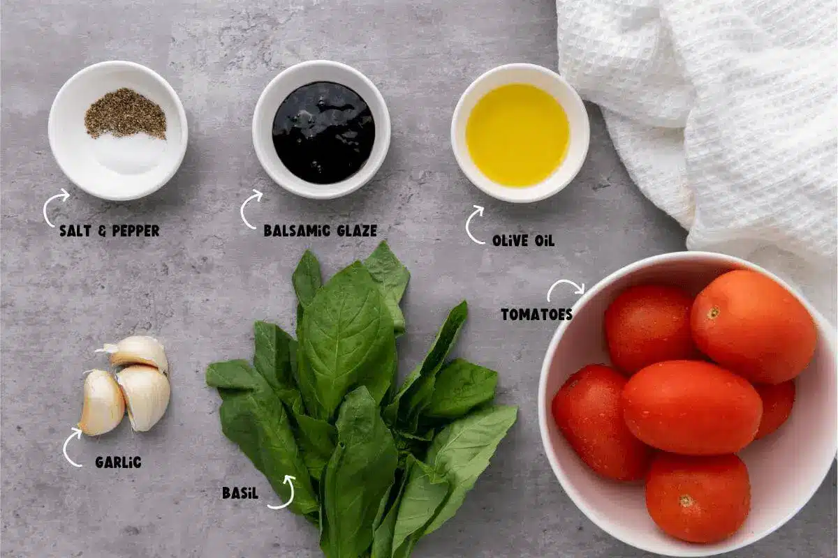 Bruschetta Ingredients_ Salt, Pepper, Balsamic Glaze, Olive Oil, Garlic, Basil, and Tomatoes sitting on a marble countertop with a white kitchen towel nearby