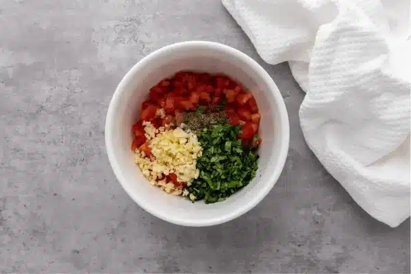 Chopped Basil, minced garlic, olive oil, salt, pepper, and diced tomatoes all in one large bowl sitting on a marble countertop with a white kitchen towel nearby