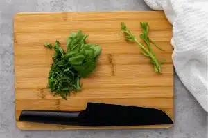 Chopped Basil on a bamboo cutting board with a black knife sitting on a marble countertop with a white kitchen towel next to the cutting board