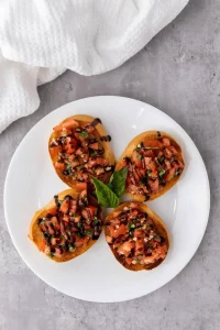 Four slices of toasted french bread topped with bruschetta and balsamic glaze on a white plate with a kitchen towel, a spoon, and a bowl of balsamic glaze nearby. nearby
