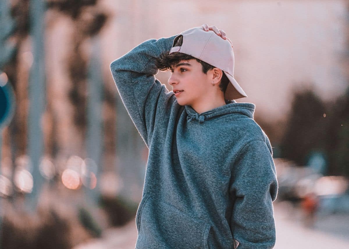 A teenage boy is wearing a tan baseball cap backwards.  He's wearing a blue/teal sweatshirt.