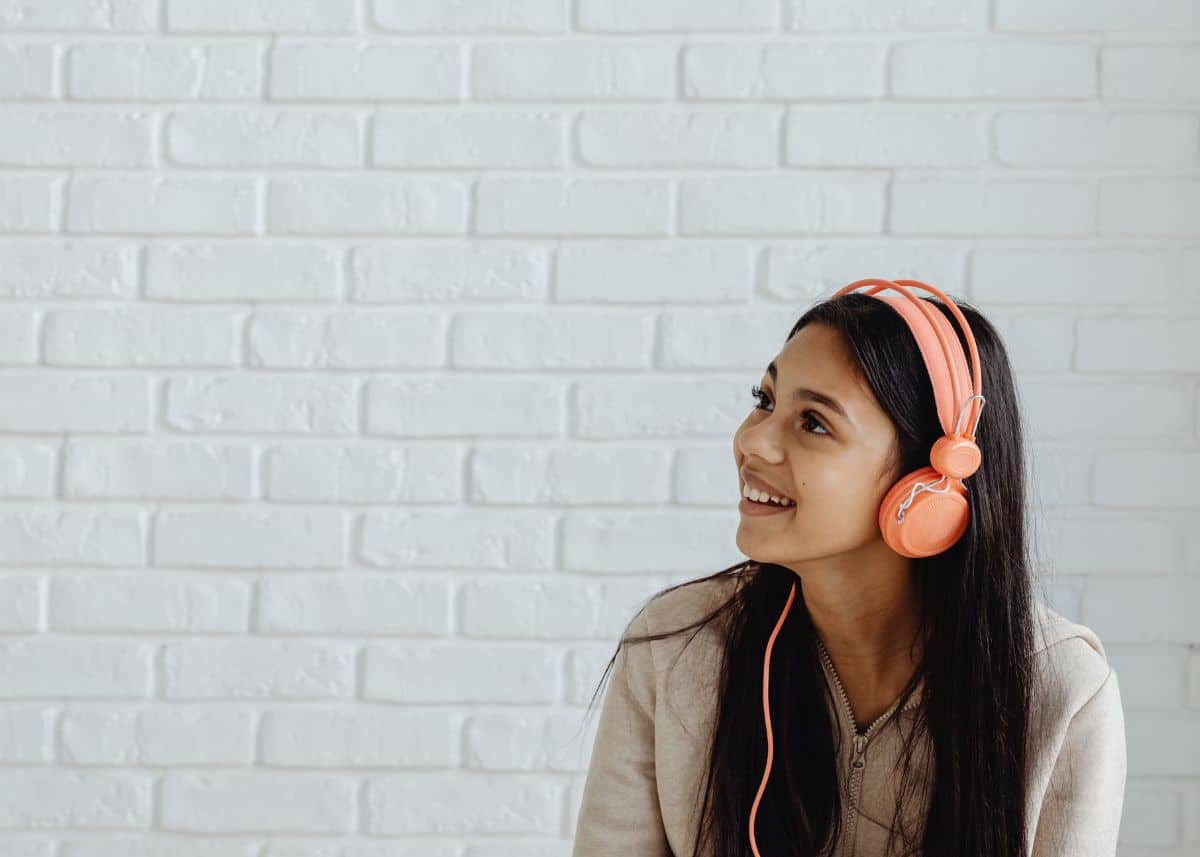 A teenage girl with long dark hair is wearing pink/coral headphones.  She's wearing a tan zip up sweatshirt.