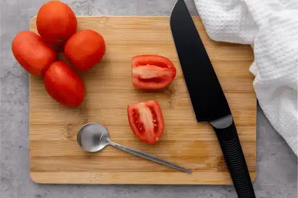 Whole and halved tomatoes on a bamboo cutting board with a black knife sitting on a marble countertop with a white kitchen towel next to the cutting board