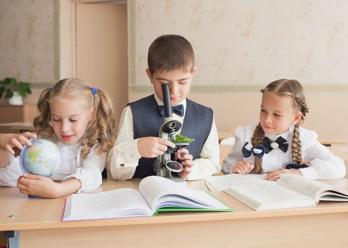 Three young students are sitting at a desk with books open.  One is using a microphone.  One is looking at a small globe.  