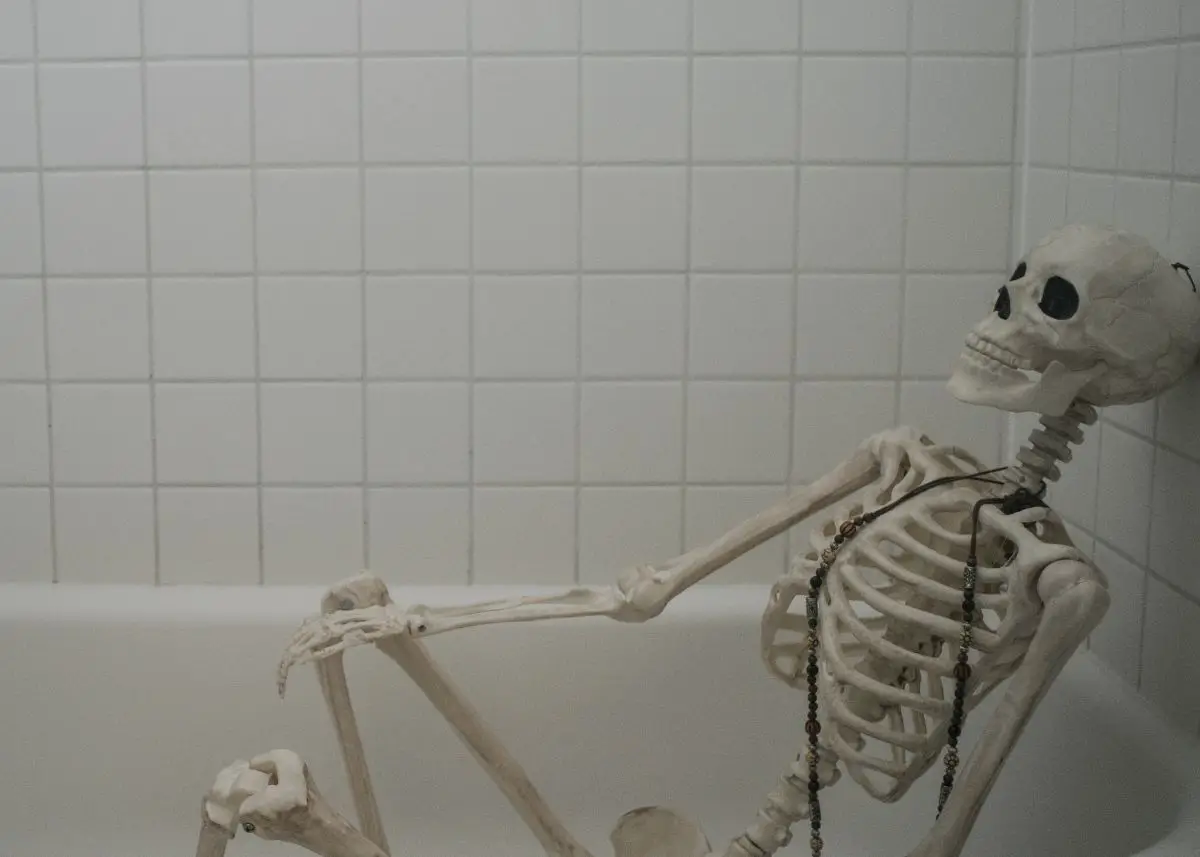 A skeleton is sitting in a bathtub.  It has a long dark necklace around its neck.  The bathtub surround is white square tiles.  