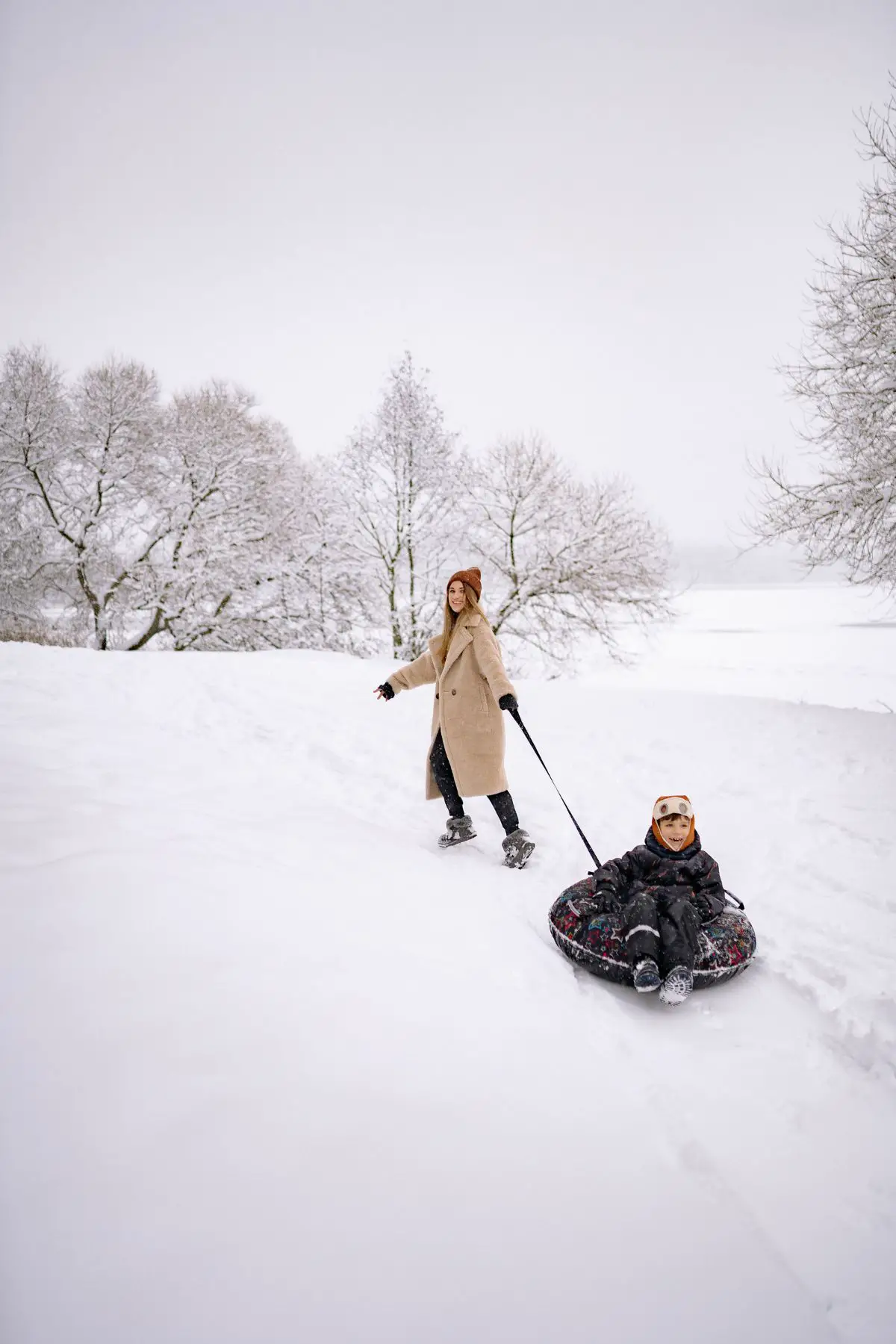 This is a photo of a mom pulling her son in a tube.  They are on a hill.  The mom is wearing a tan peat coat and the son is wearing a black snow suit.  