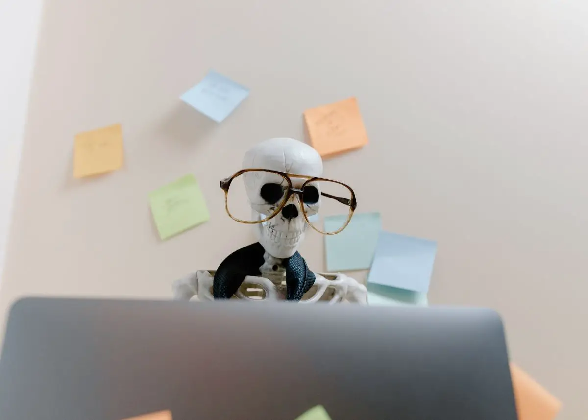 This is a photo of a skeleton sitting behind a computer.  It's wearing giant glasses and a tie.  There are pastel colored post it notes on the wall in the background.