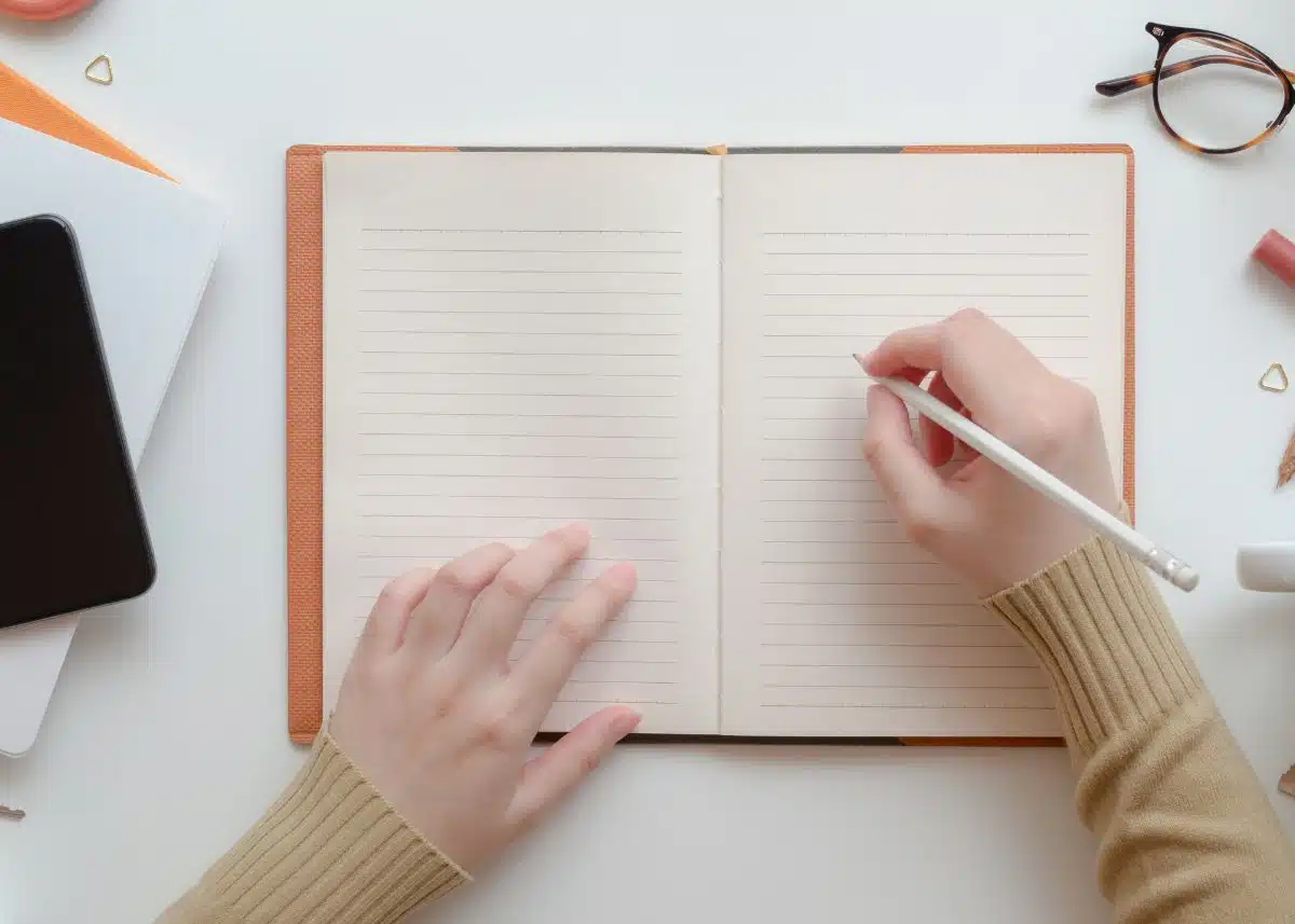 This is a picture of someone holding a pencil writing in a notebook.  Glasses and a tablet are next to the journal.