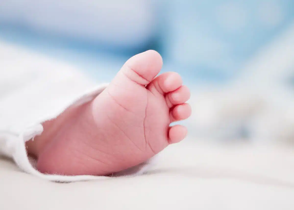 The is a closeup of the bottom of a baby's left foot.  There's a blue background.  The baby is wearing white pants.