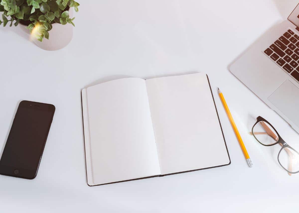 This is an overhead view of a journal lying open on a white desk.  There's a pencil, glasses, phone, laptop, and plant also in the photo.  