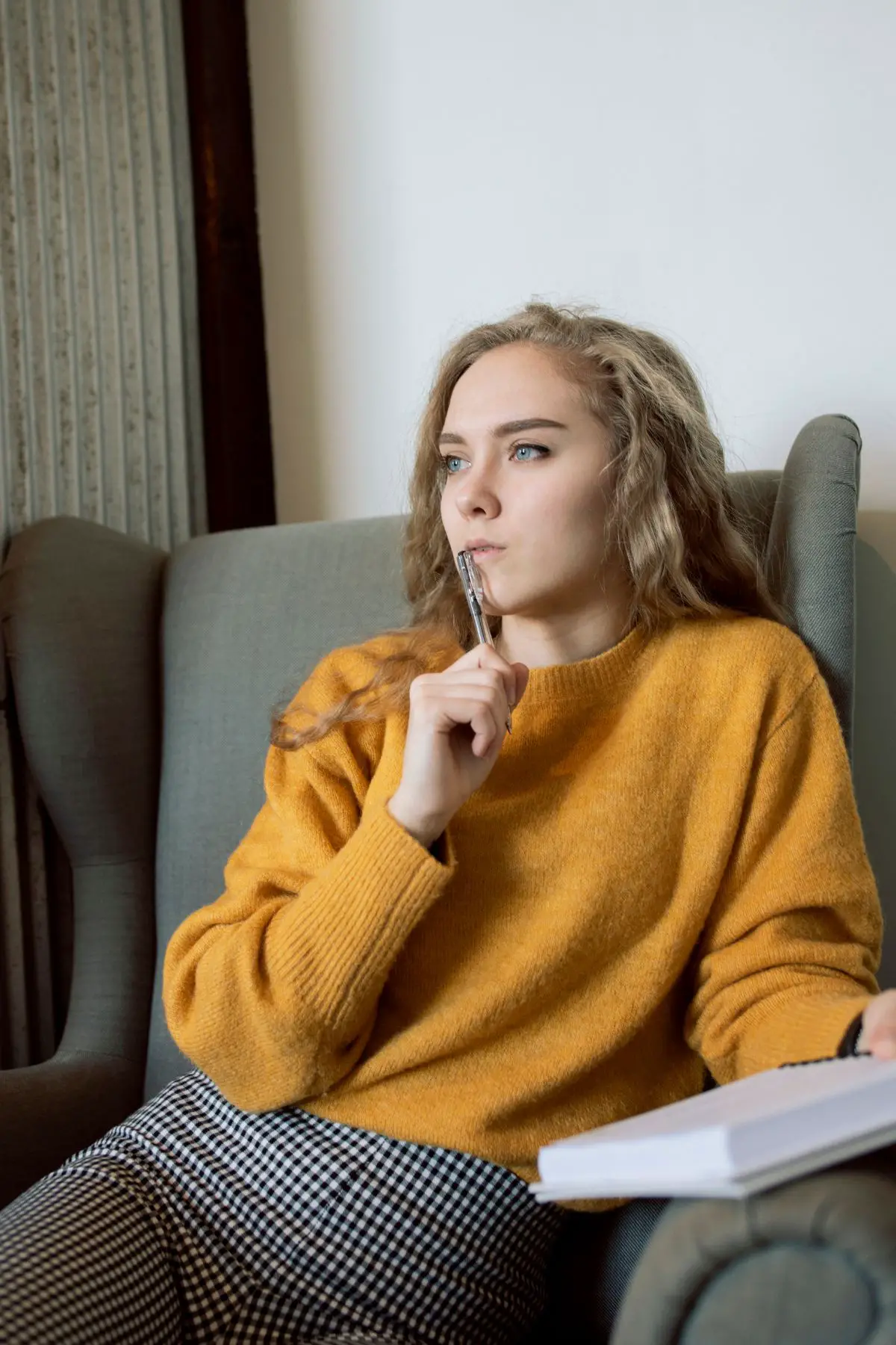 This is a photo of a teenage girl sitting in a chair.  She's holding a pen up to her mouth and she's thinking.  She's holding a notebook too.