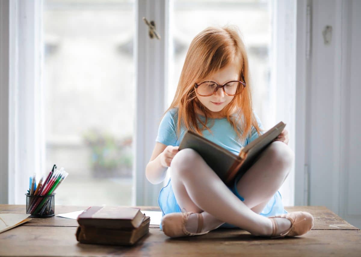 A little girl is reading a book while sitting on a desk at home.  There are two books next to her.  She has red hair, she has glasses, and she's wearing a blue dress and ballet slippers.